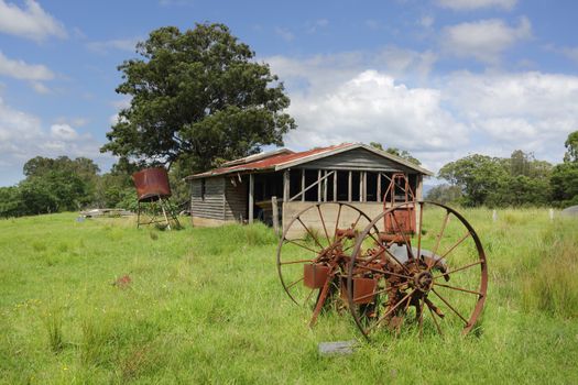 Old farm shed or barn, with a rusty water tank and cart wheels lie in overgrown grass in Benandarah NSW Australia