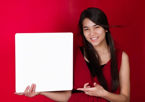 Beautiful biracial teen girl holding up square white sign, pointing to it on red background