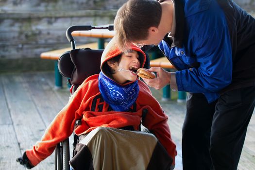 Father feeding disabled son a hamburger in wheelchair. Child has cerebral palsy