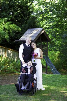 Interracial bride and groom standing with her disabled little boy in wheelchair for wedding pictures outdoors