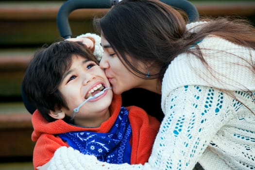 Big sister kissing disabled little brother seated in wheelchair on cheek