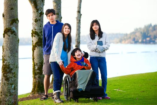 Disabled little boy in wheelchair surrounded by brother and sisters at lakeside. Child has cerebral palsy and children are all biracial