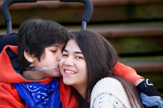 Disabled little boy kissing his big sister on cheek while seated in wheelchair