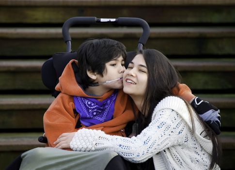 Disabled little boy kissing his big sister on cheek while seated in wheelchair