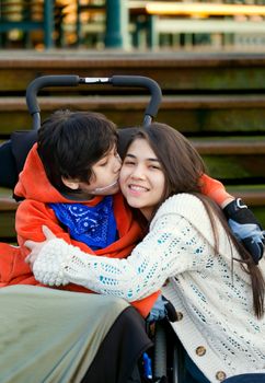 Disabled little boy kissing his big sister on cheek while seated in wheelchair