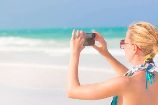 Blonde caucasian woman taking photo of blue tropical beach. Beautiful caucasian model  wearing turquoise swimsuit and colorful scarf on vacations on picture perfect Paje beach, Zanzibar, Tanzania.