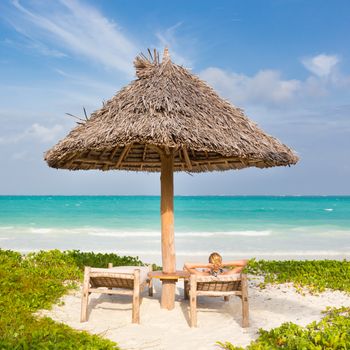 Women sunbathing on dack chair under wooden umbrella on stunning tropical beach. Turquoise blue lagoon of Paje beach, Zanzibar, Tanzania in the background.