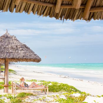 Women sunbathing on dack chair under wooden umbrella on stunning tropical beach. Kiteboarders turquoise blue lagoon of Paje beach, Zanzibar, Tanzania in the background.