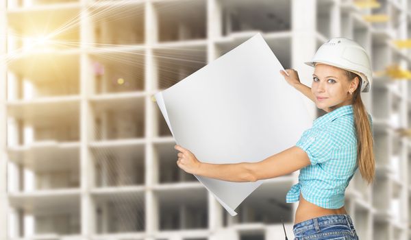 Woman in hard hat holding blank drawing sheet in front of building under construction, looking, at camera, smiling