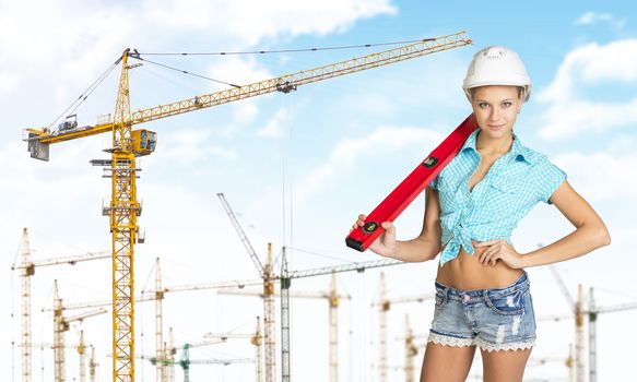 Woman in hard hat with large builders level on her shoulder, looking at camera. Sky, clouds and tower cranes as backdrop
