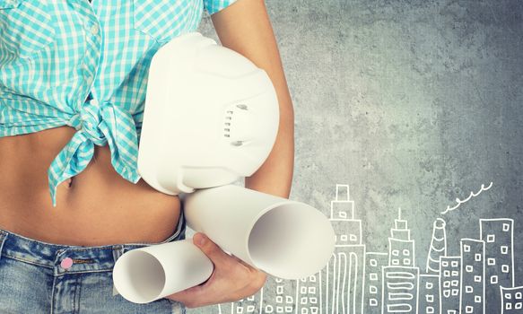 Close-up of woman holding hard hat and drawing rolls, against stone wall with sketch of city on it 