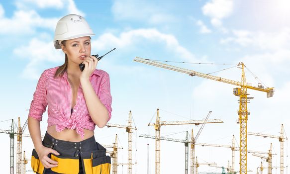 Woman in hard hat and tool belt talking on walkie talkie, looking at camera. Sky, clouds and tower cranes as backdrop