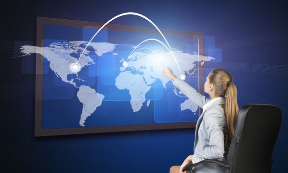 Businesswoman operating touch screen interface featuring world map with flight paths or trade routes or communication between countries and cities and on blue background
