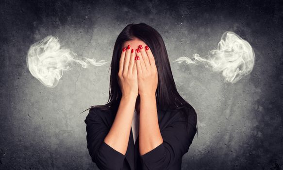 Close-up portrait of businesswoman hiding her face in her hands, with smoke from her ears. Raw concrete wall as backdrop