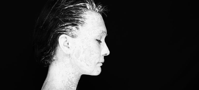 Black and white portrait of pretty young woman with clay on face on black background