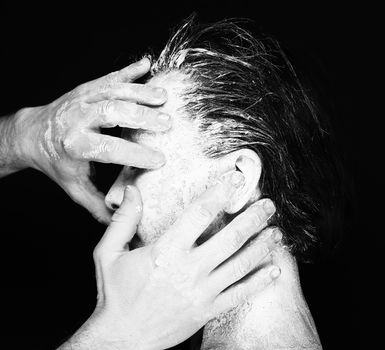 Black and white portrait of human hands working with woman with clay on face on black background