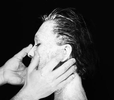 Black and white portrait of human hands working with woman with clay on face on black background