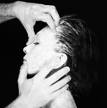 Black and white portrait of human hands working with woman with clay on face on black background