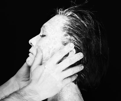 Black and white portrait of human hands working with woman with clay on face on black background