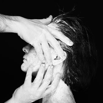 Black and white portrait of human hands working with woman with clay on face on black background