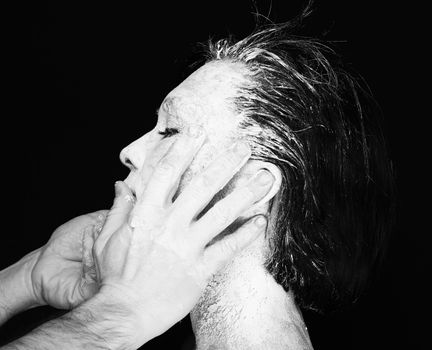 Black and white portrait of human hands working with woman with clay on face on black background