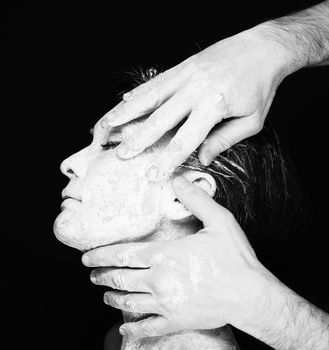 Black and white portrait of human hands working with woman with clay on face on black background