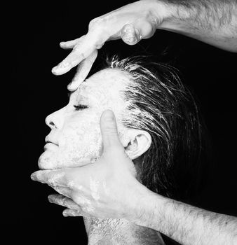 Black and white portrait of human hands working with woman with clay on face on black background