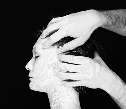 Black and white portrait of human hands working with woman with clay on face on black background