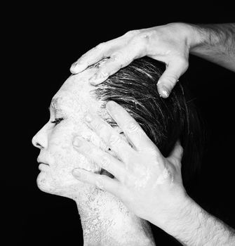 Black and white portrait of human hands working with woman with clay on face on black background