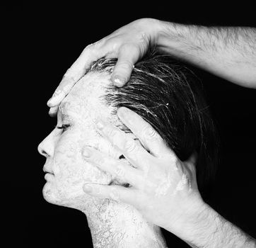 Black and white portrait of human hands working with woman with clay on face on black background