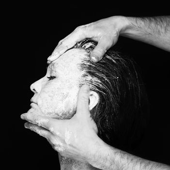 Black and white portrait of human hands working with woman with clay on face on black background