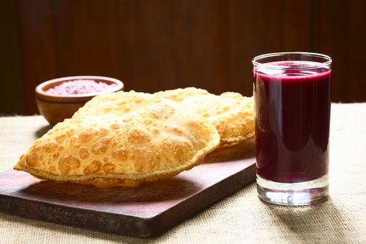 Traditional Bolivian snack called Pastel (deep-fried pastry filled with cheese) served with Api, a purple corn beverage on wooden board, photographed with natural light (Selective Focus, Focus on the front of the pastry and the glass)   