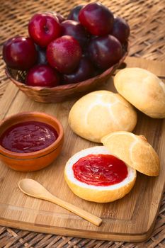 Plum jam spread on bun with plums in basket on wooden board photographed with natural light (Selective Focus, Focus on the front of the jam on the bun)