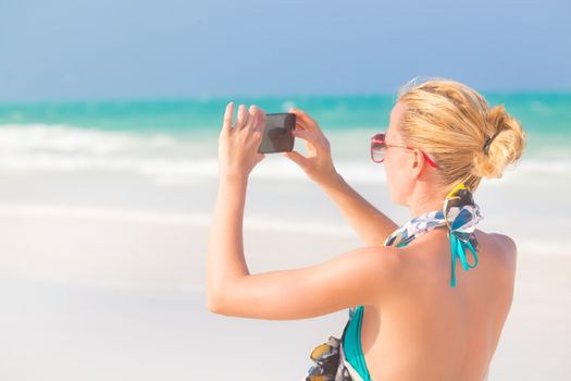 Blonde caucasian woman taking photo of blue tropical beach. Beautiful caucasian model  wearing turquoise swimsuit and colorful scarf on vacations on picture perfect Paje beach, Zanzibar, Tanzania.