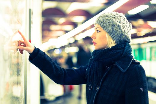 Casually dressed woman wearing winter coat, orientating herself with public transport map panel, pointing on her final destination. Urban transport.