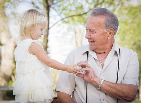 Cute Baby Girl Handing Easter Egg to Grandfather Outside at the Park.