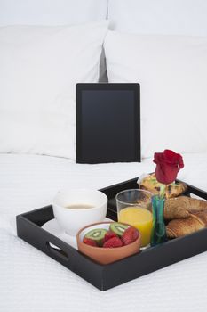 white quilt bed breakfast black tray croissants orange juice strawberry kiwi cupcake red rose flower and digital tablet blank screen