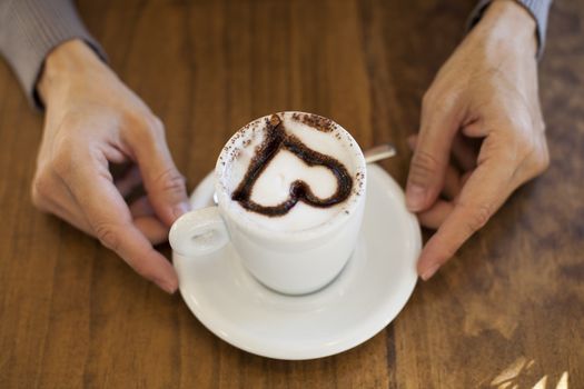 white small cup cappuccino coffee with chocolate heart in woman hands on light brown wooden table