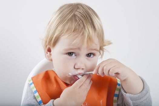 blonde caucasian baby seventeen month age orange bib grey sweater in white high chair licking spoon yogurt