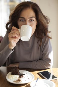 woman drinking white small cup cappuccino coffee looking at camera with chocolate cake piece spoon and mobile phone blank screen on light brown wooden table