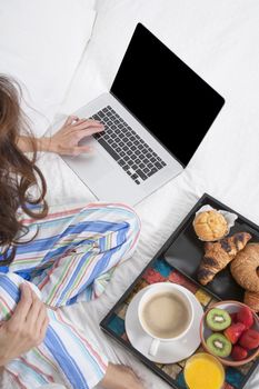 woman green shirt striped pajama pants sitting on white bed typing keyboard laptop with breakfast tray croissants orange juice strawberry kiwi cupcake red rose flower