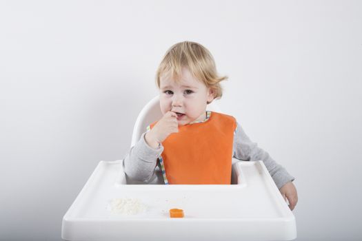 blonde caucasian baby seventeen month age orange bib grey sweater eating meal in white high-chair looking at camera