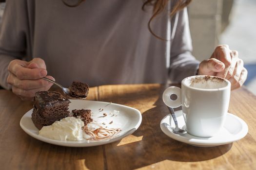 woman hands taking chocolate cake piece with spoon and white small cup cappuccino coffee on light brown wooden table