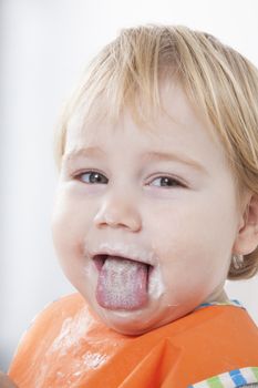 blonde caucasian baby seventeen month age orange bib on white background sticking out tongue with yogurt