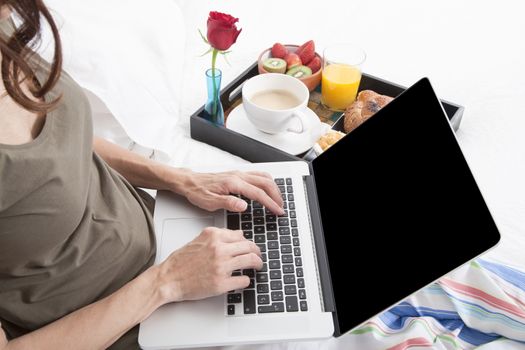 woman green shirt striped pajama pants sitting on white bed typing keyboard laptop with breakfast tray croissants orange juice strawberry kiwi cupcake red rose flower