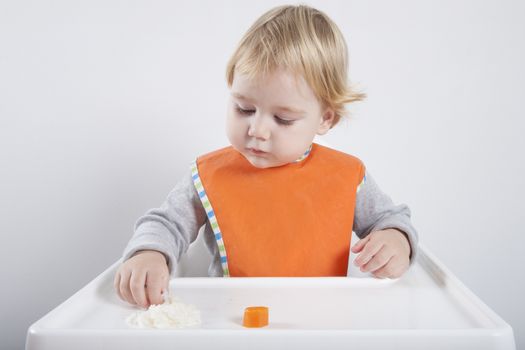blonde caucasian baby seventeen month age orange bib grey sweater eating meal with her hand in white high-chair