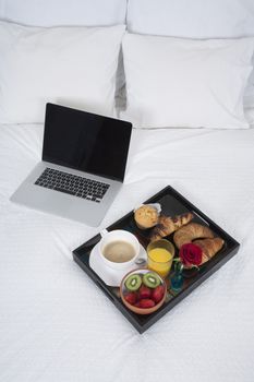 white quilt bed breakfast black tray croissants orange juice strawberry kiwi cupcake red rose flower and silver laptop blank screen