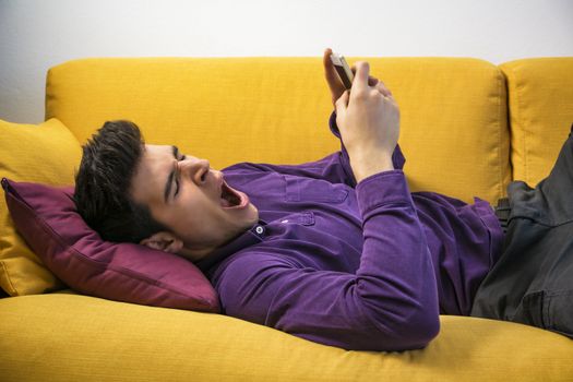 Attractive young man using cell phone and yawning, tired, while laying on couch, with serious expression