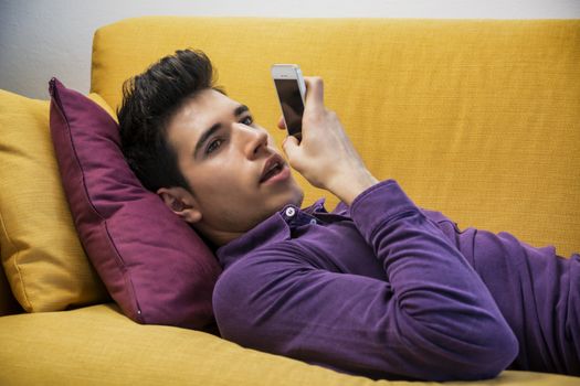 Attractive young man recording voice message on cell phone while laying on couch, with serious expression