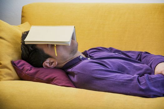 Over-worked, tired young man at home sleeping instead of working or studying, resting with head covered by open book